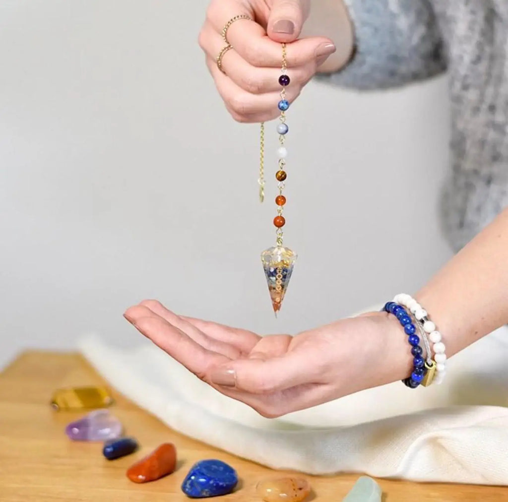 Pêndulo de orgonite dos 7 chakras a balançar sobre a mão aberta, com pedras soltas na mesa