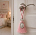 Pingente decorativo com medalha milagrosa pendurado na maçaneta de uma porta branca, quarto ao fundo